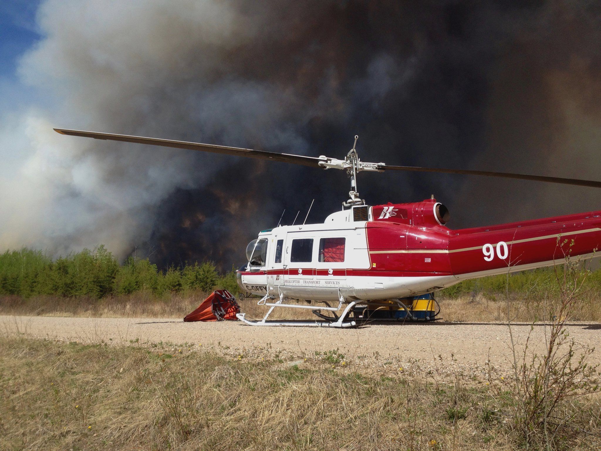 HTS Bell 205 on a gravel pad with wildfire smoke rising behind it