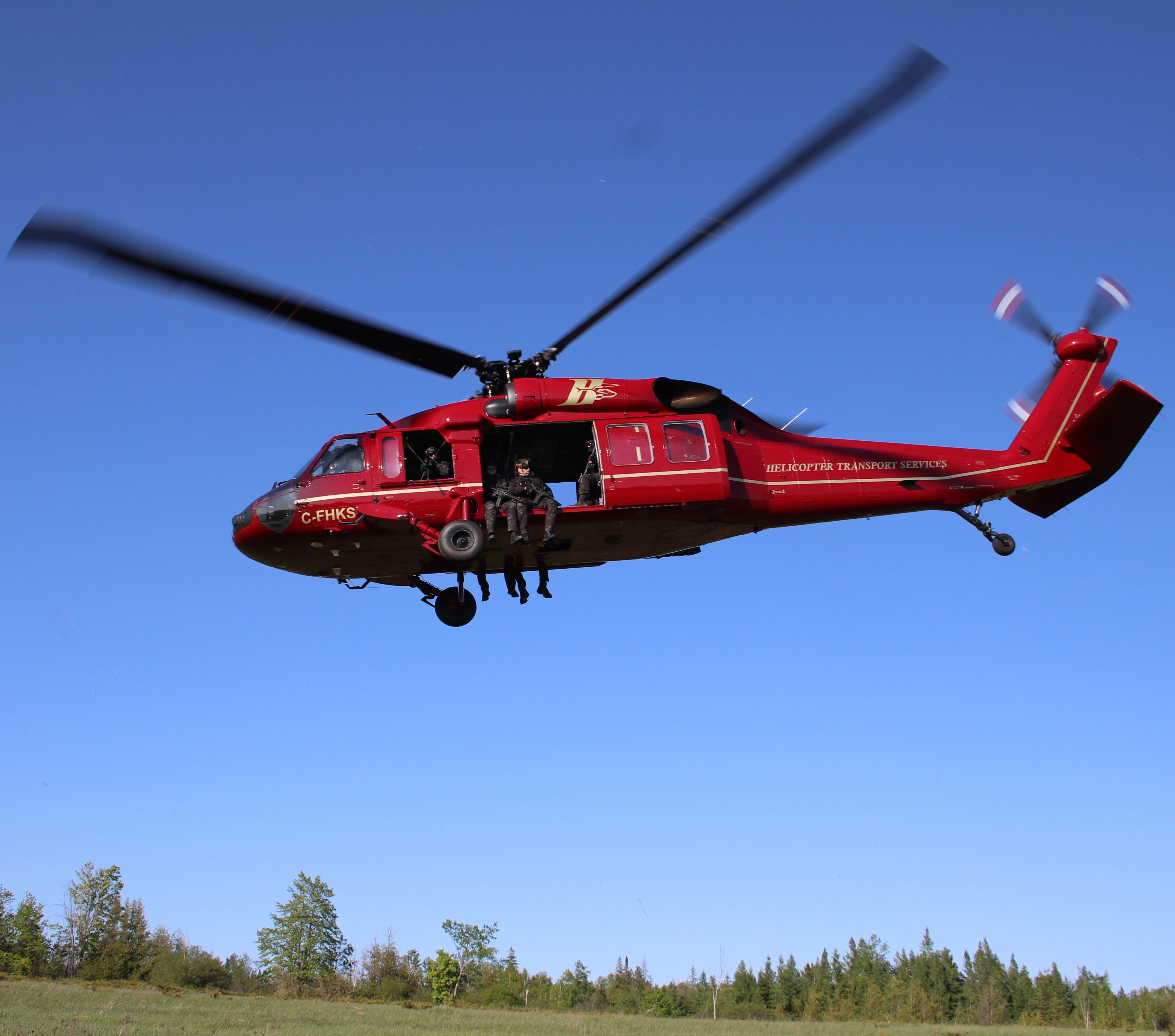 HTS Black Hawk in flight against a clear sky, a crew member seated in the open door