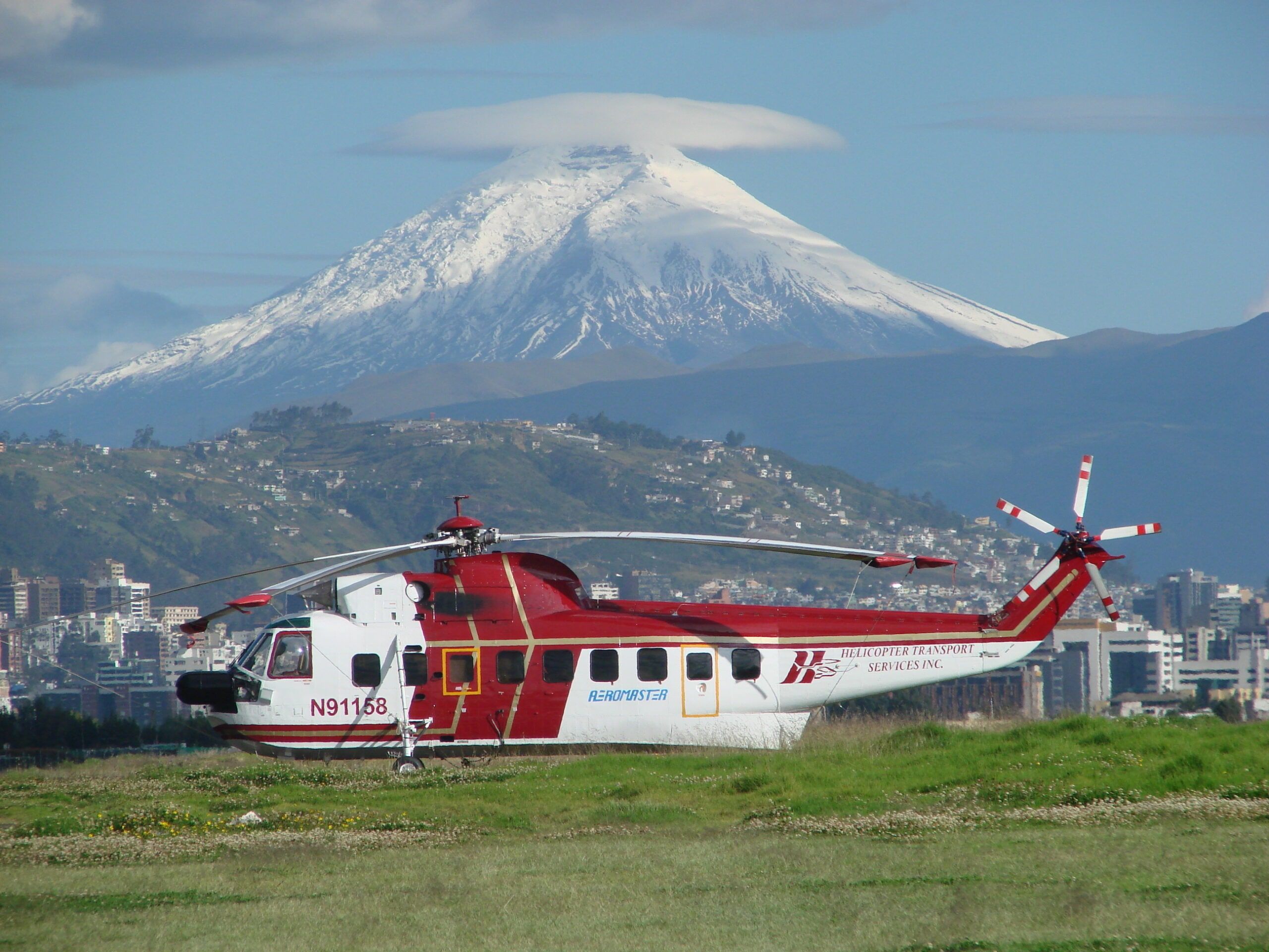 HTS S-61 parked on a hillside field above a city, a snow-capped volcano behind