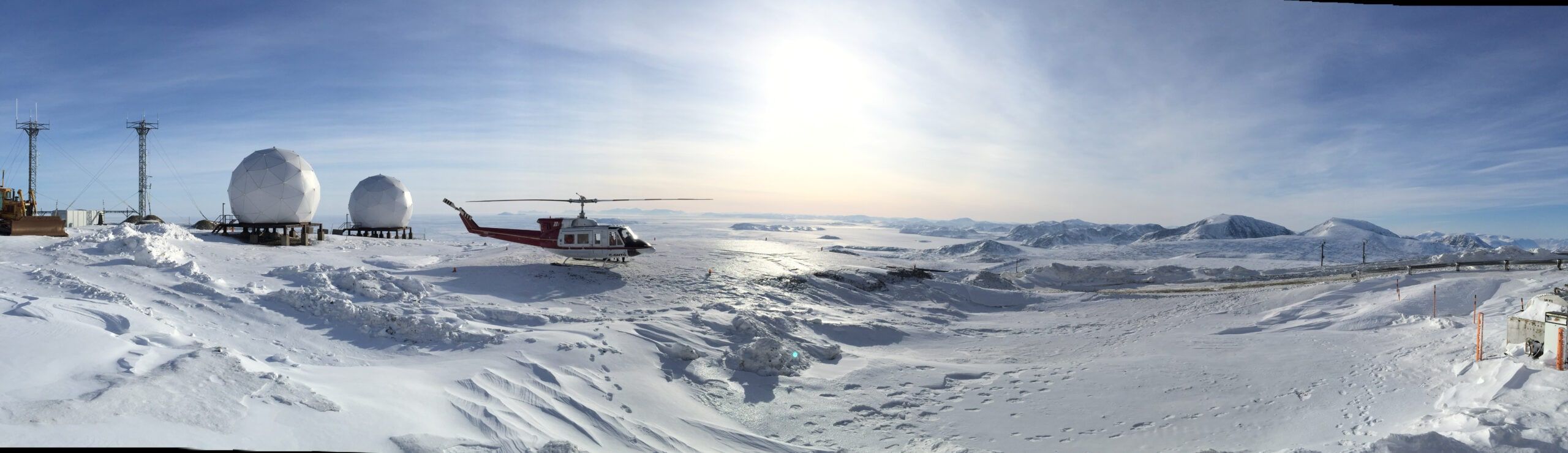 A helicopter flying low over snow and sea ice under a bright Arctic sun