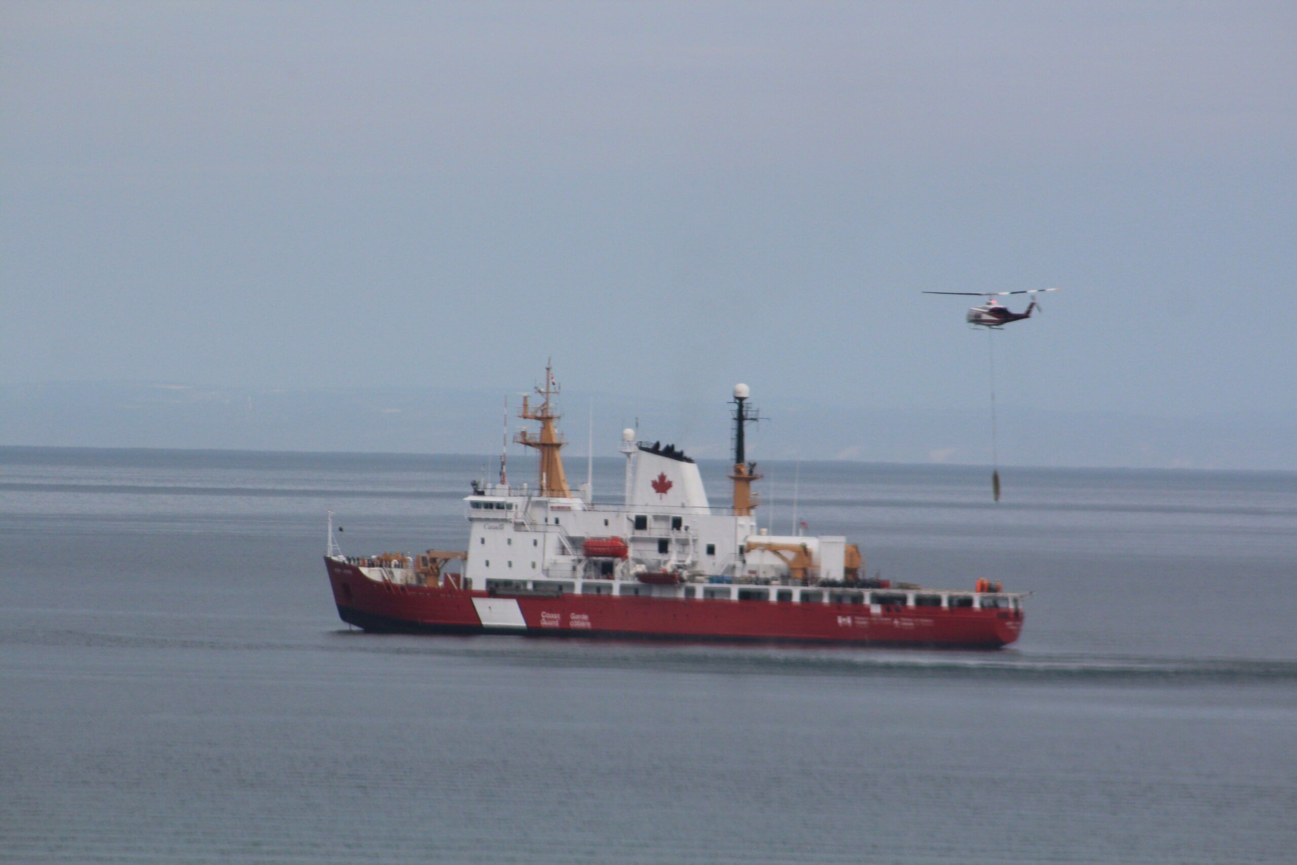 Red and white coast guard icebreaker at sea with a helicopter flying above it