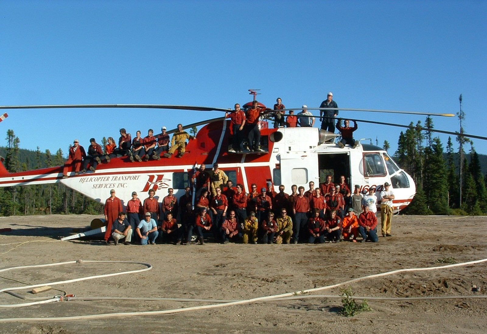 A large HTS crew gathered in front of a red and white S-61 on a gravel pad