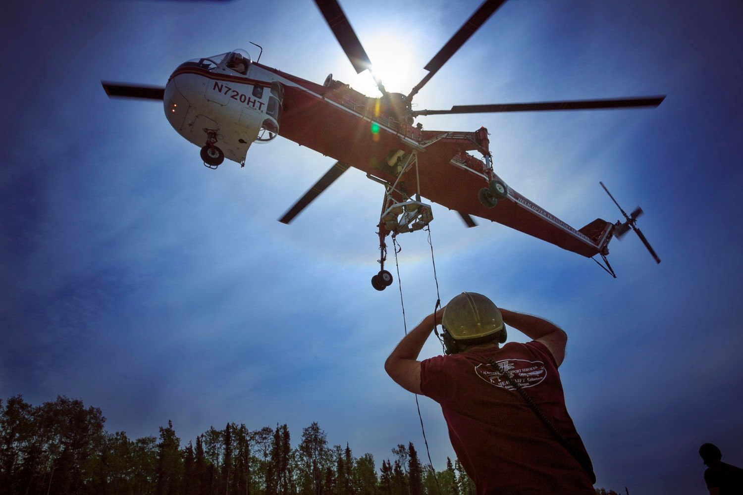 A crew member in a hard hat looks up at a Skycrane hovering against the sun