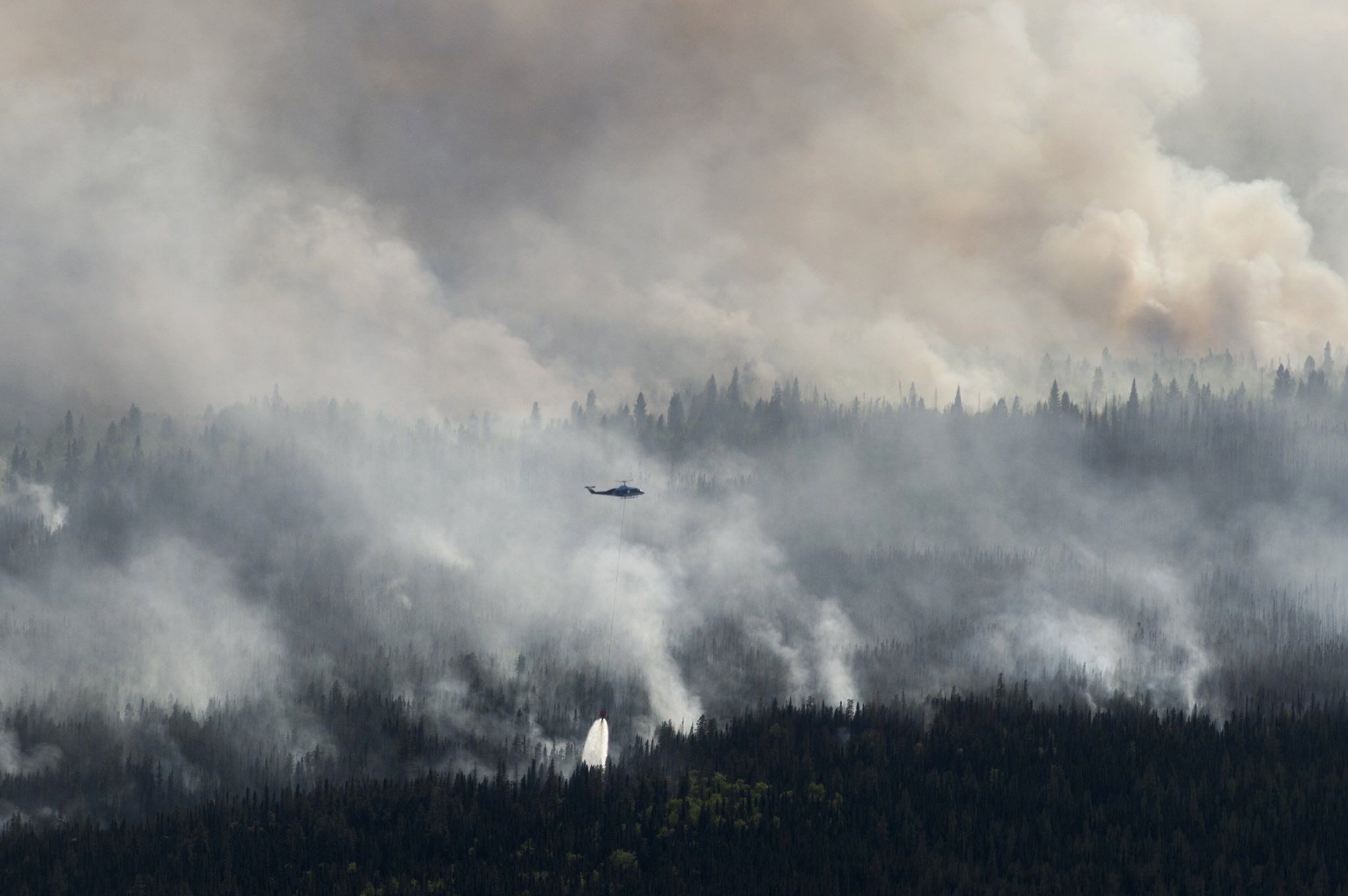 A helicopter working through heavy wildfire smoke above a burning forest