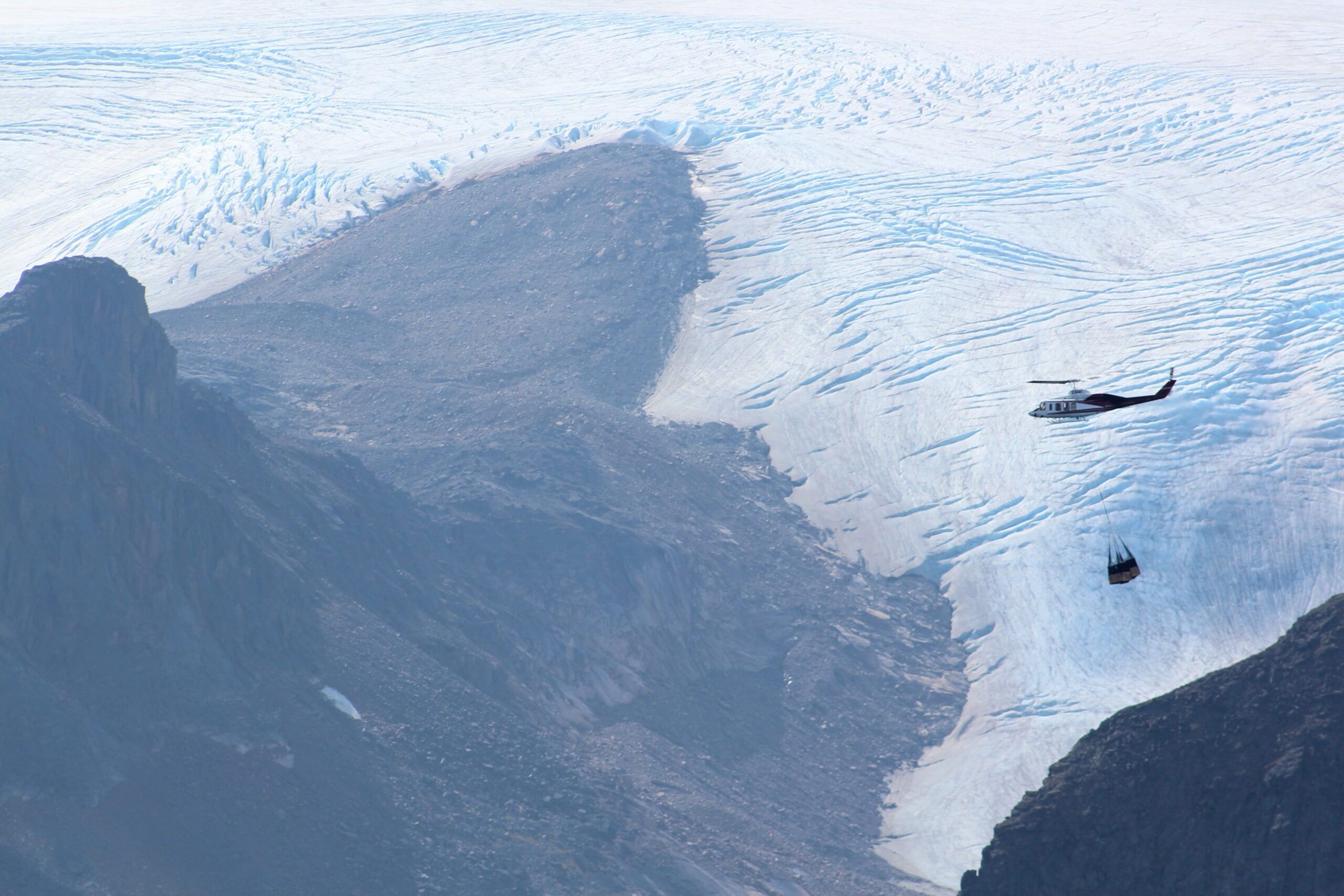 A helicopter carrying a load on a long line above a glacier between dark mountains