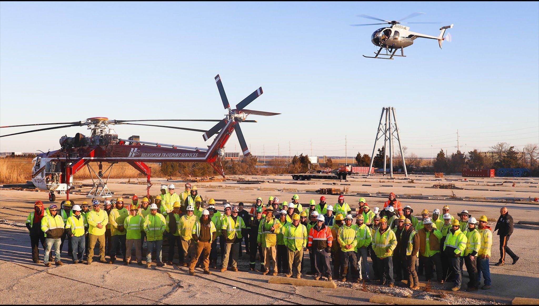 The HTS team in high-visibility vests lined up on a job site in front of a Skycrane, with a light helicopter flying overhead