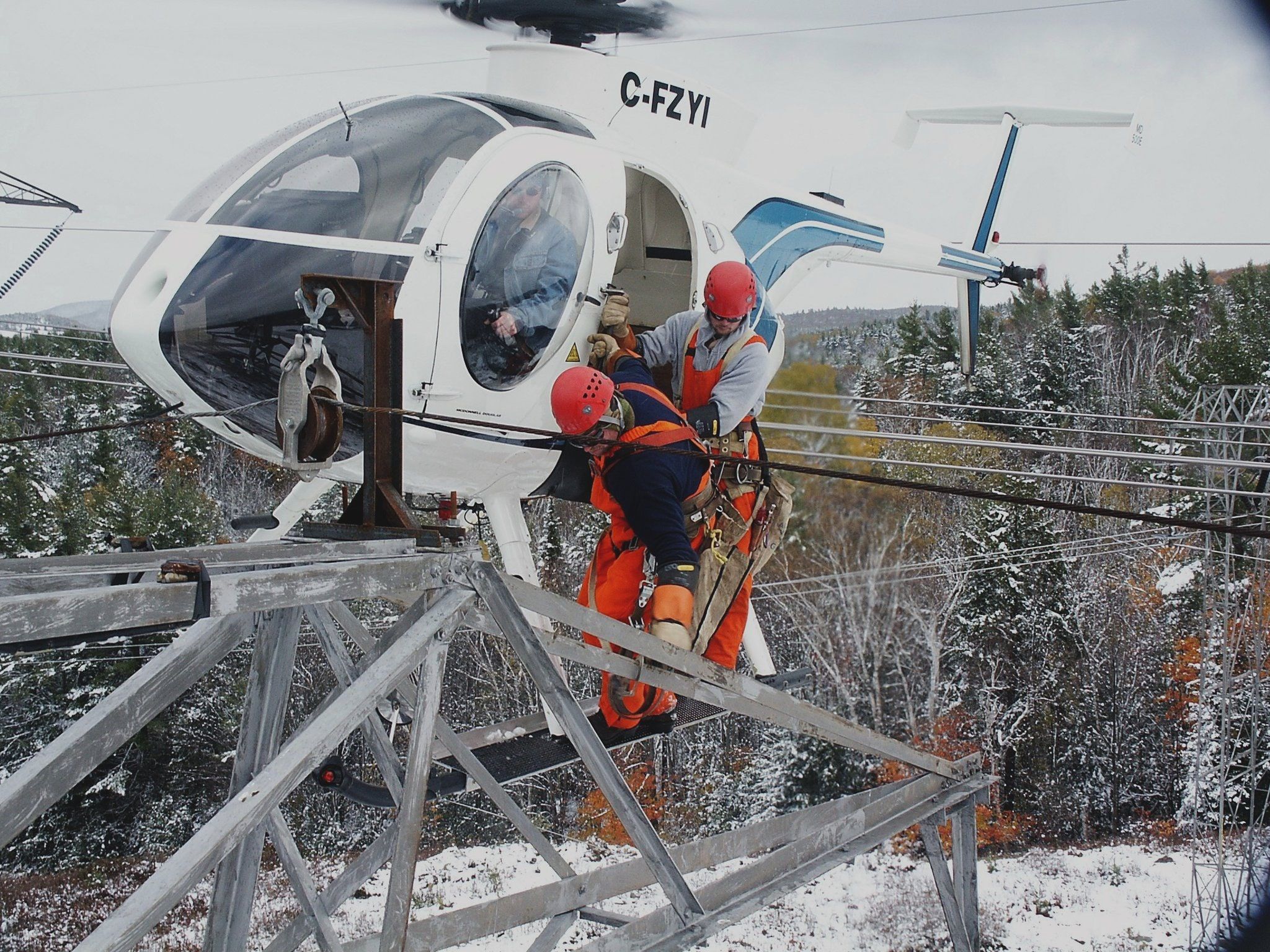 Two linemen in orange working from the skid of a light helicopter beside a steel tower in winter