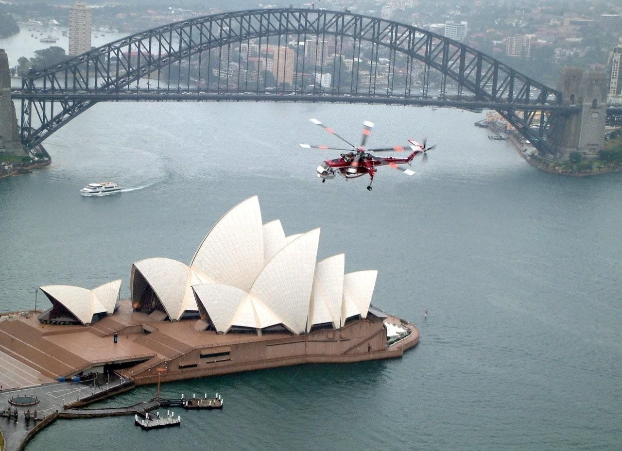 A Skycrane flying over Sydney Harbour beside the Opera House and the Harbour Bridge