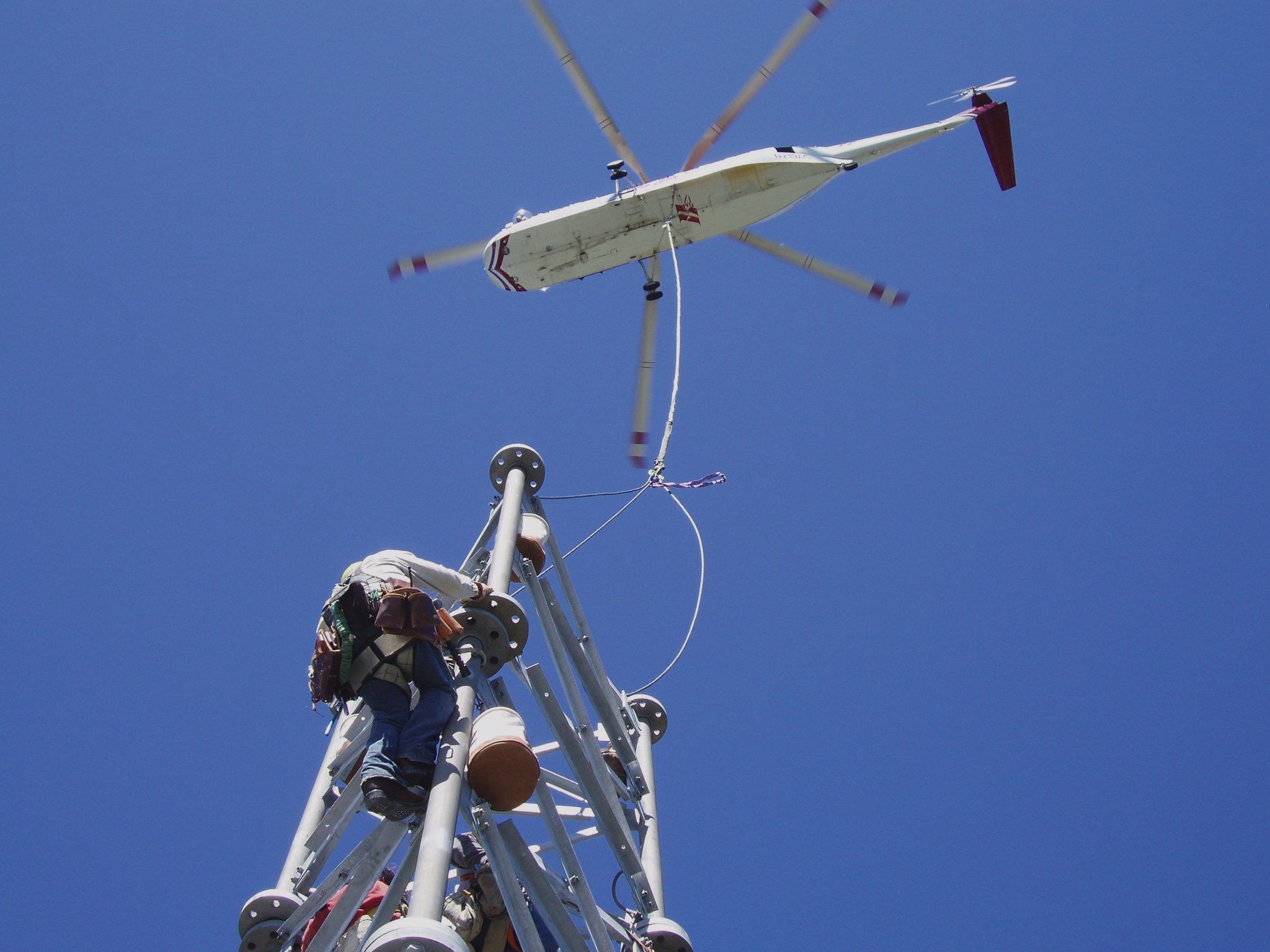 A lineman standing on top of a steel tower, guiding a load on a long line beneath a helicopter